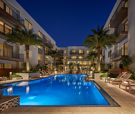 Dusk pool view with lights, pool lounge chairs, palm trees, and views of community in background