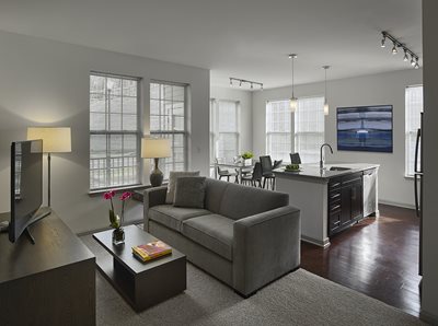 Fully furnished living room with gray sofa, dark wood tables and kitchen in background