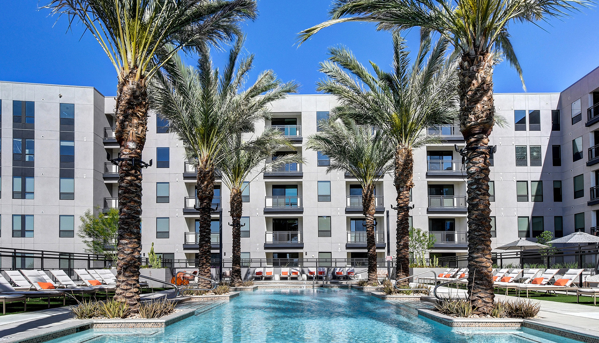 Pool in daytime with palm trees, lounge chairs with orange pillows, gray pool umbrellas and views of community