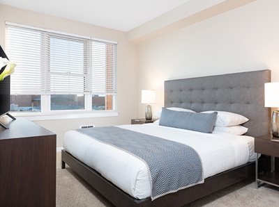 Bedroom with king-sized bed, gray headboard, white linens and pillows, gray throw blanket and oak dresser with tv