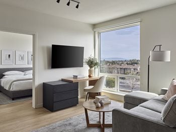 Living Room with gray sofa, white club chair and circular dining table, large windows leading to balcony, tv on oak stand