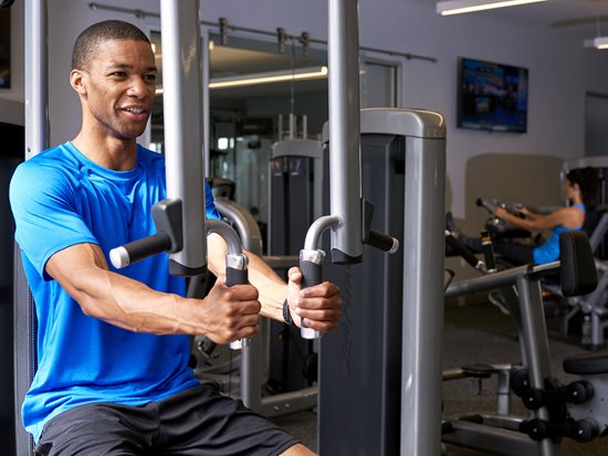 Man in blue shirt using fitness machine with woman in light blue shirt behind him