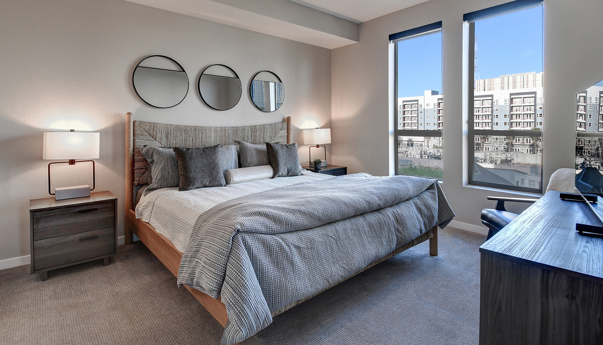 Bedroom with king-sized bed, wood bed frame and headboard, white and gray blankets and pillows, and views of community through windows