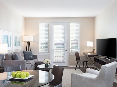 Living Room with gray sofa, white club chair and circular dining table, large windows leading to balcony, tv on oak stand