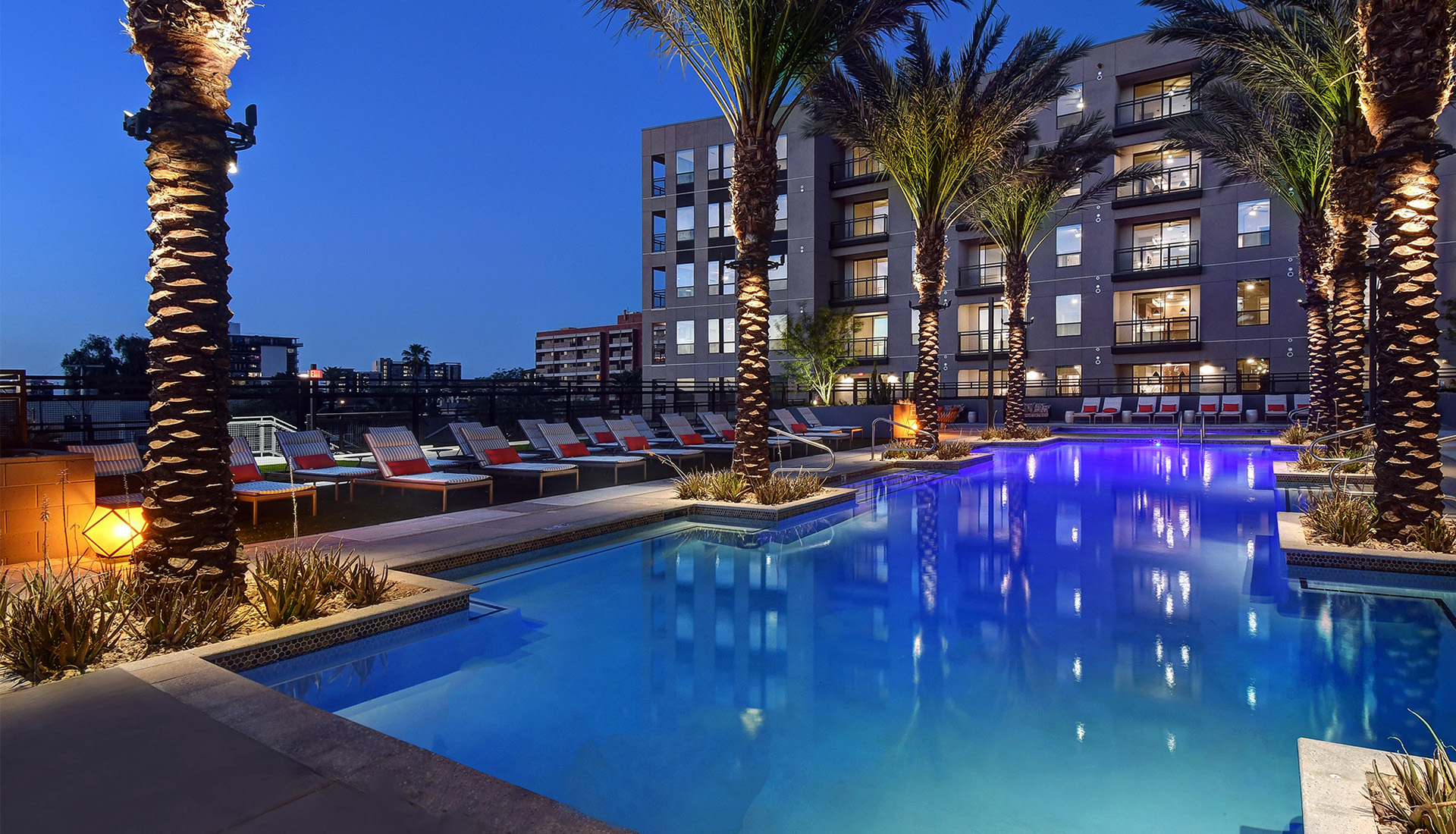 Resort style pool at dusk with palm trees, lanterns and lounge chairs with red pillows. views of community in backgroun